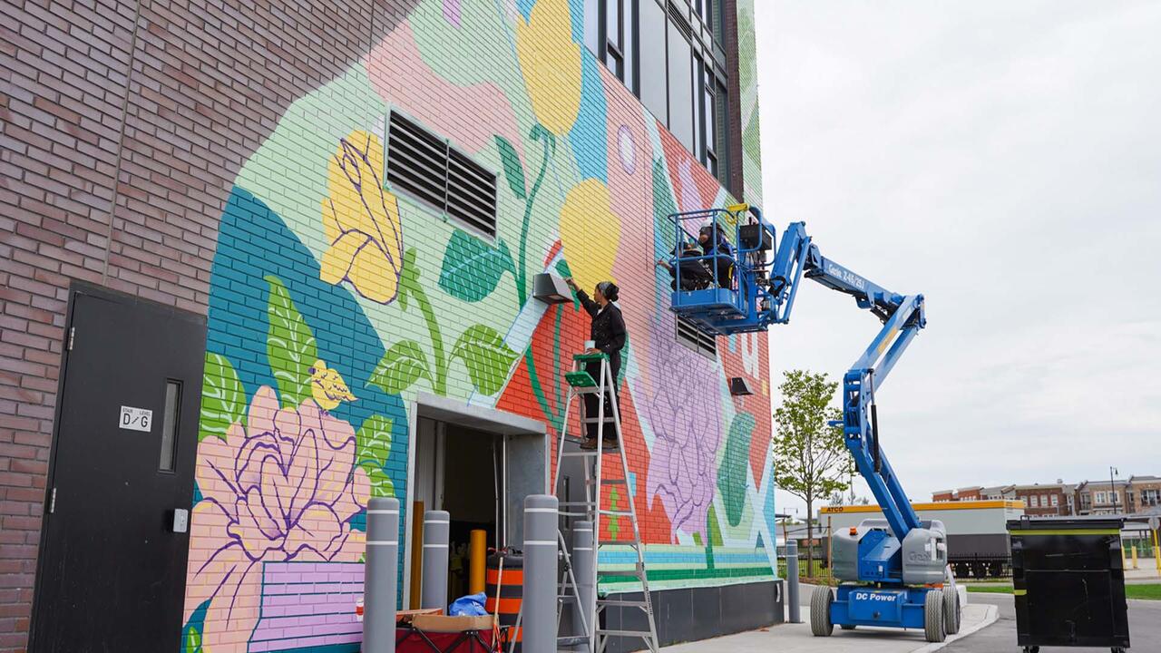 Two students - one on a ladder and one in a construction lift - painting a bright coloured mural on the side of a building